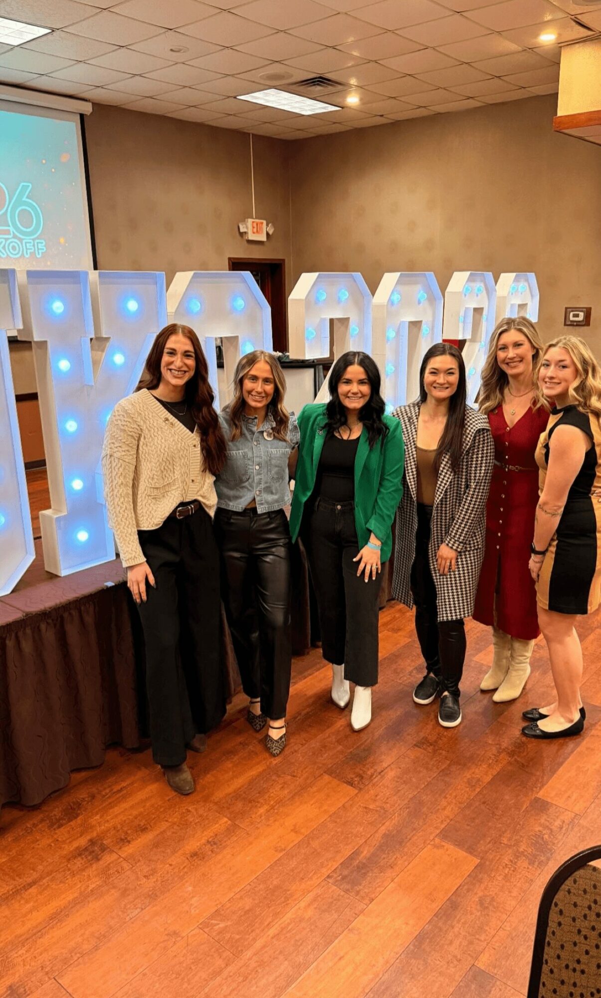 Six women stand together indoors, smiling, in front of large illuminated letters spelling 'DREAM'.