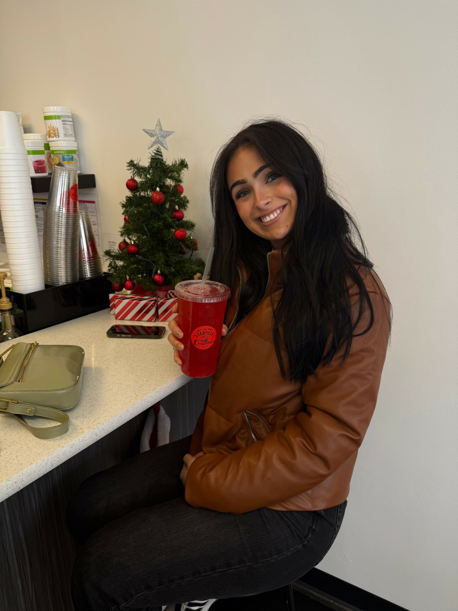 Young woman with long dark hair smiling, sitting at a counter with a small decorated Christmas tree and red cup.
