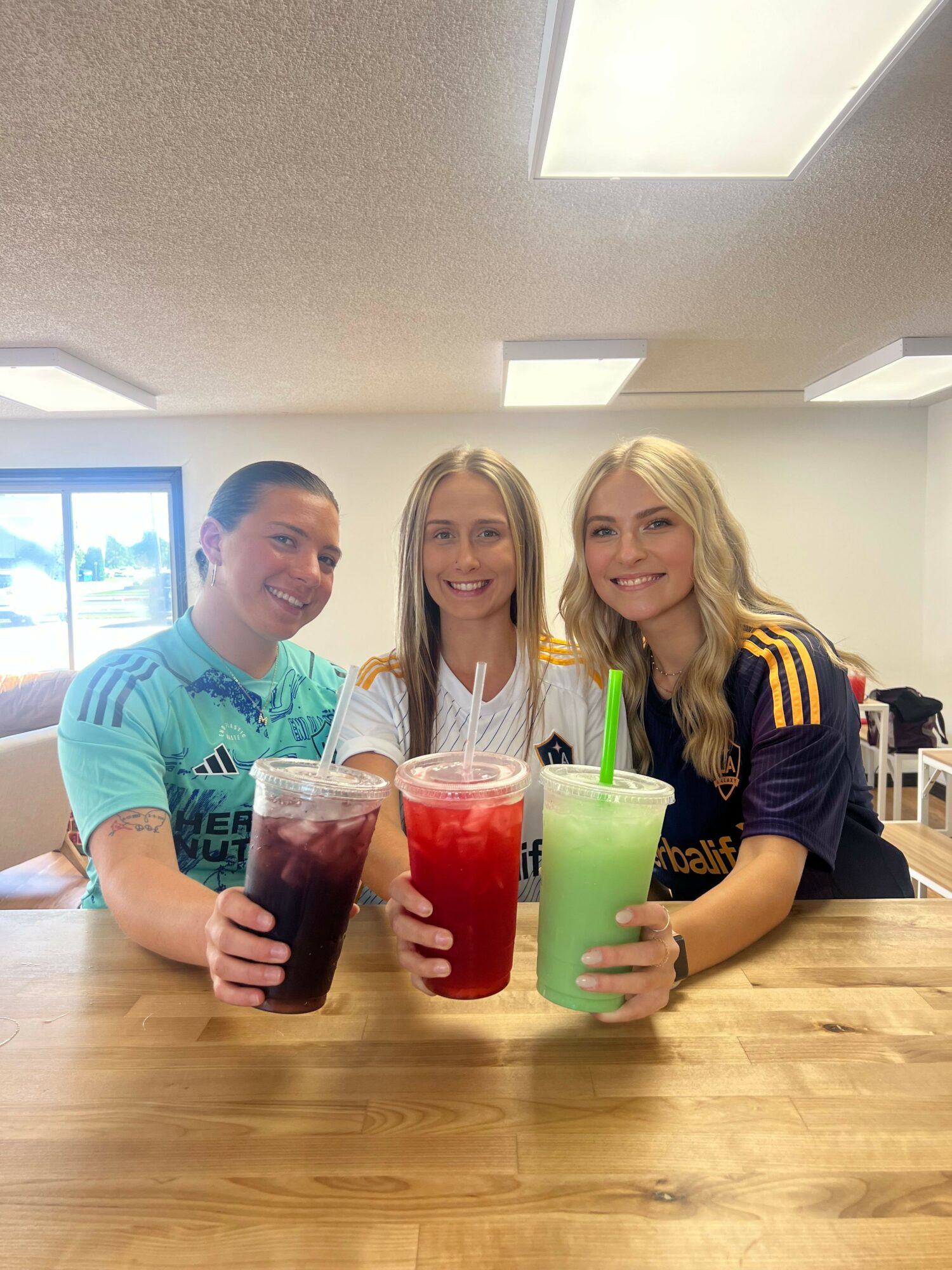 Three smiling women holding colorful drinks at a table indoors, with a window and ceiling lights visible.