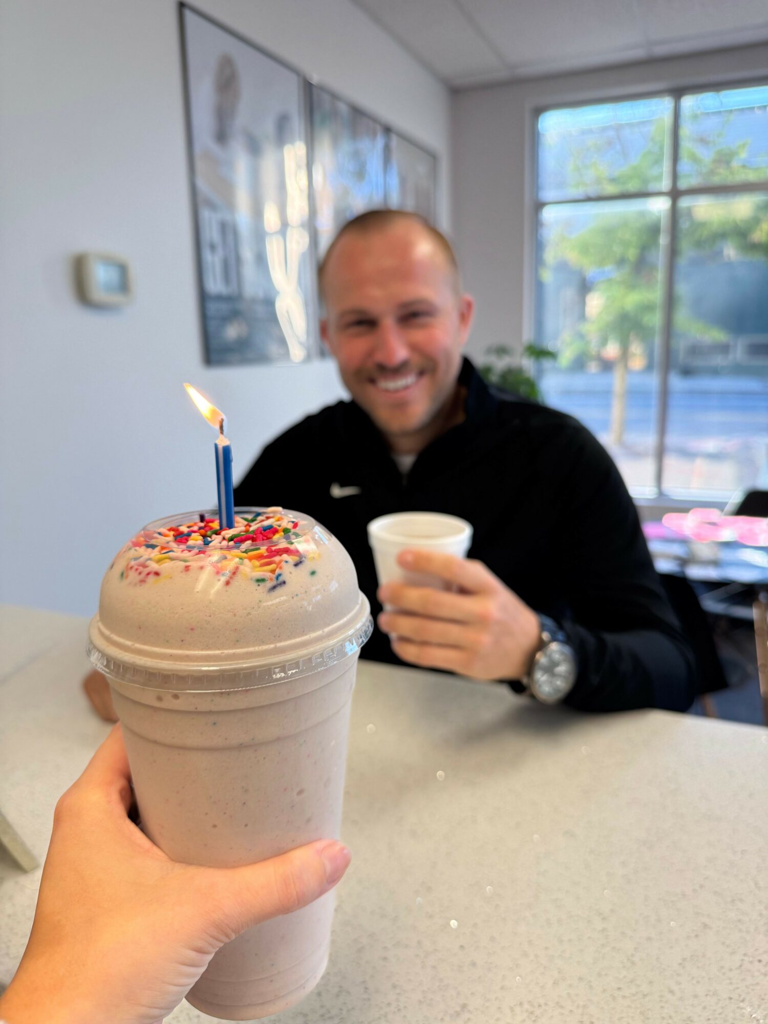 Person holding a large milkshake with a lit birthday candle and colorful sprinkles, smiling at a man in the background.