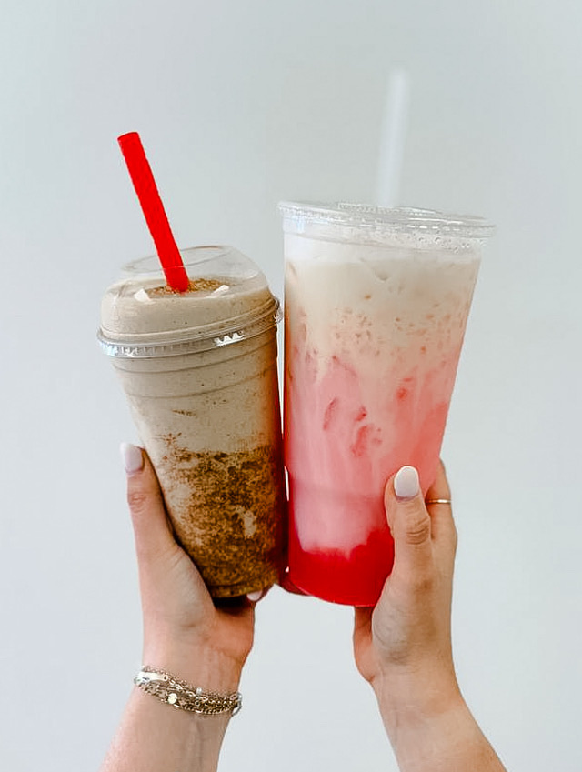 Two hands holding different colorful beverages with straws, one brown and one pink, against a plain background.