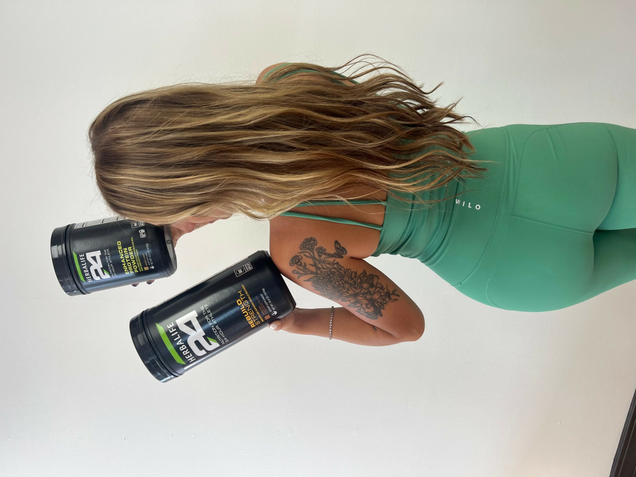 Woman with long hair holding two black cans, wearing green athletic clothing, standing sideways against a plain background.