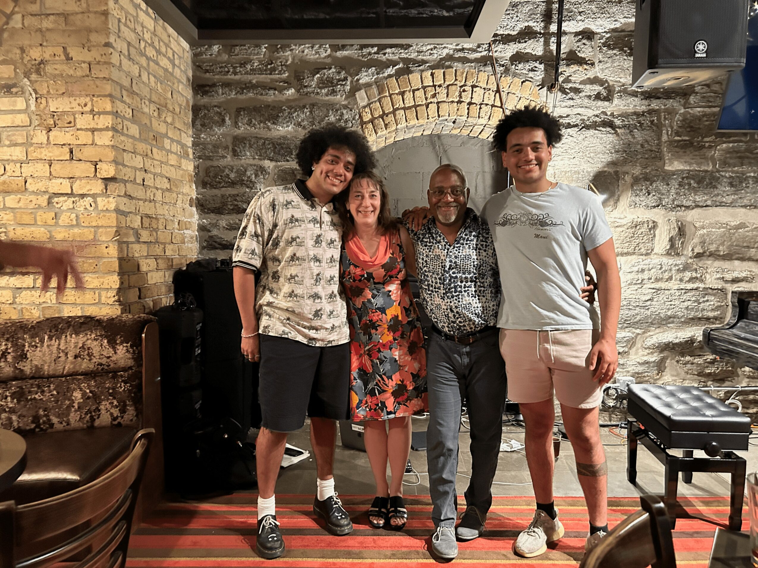 Four people standing together in front of a brick and stone wall, smiling, with a small table nearby.
