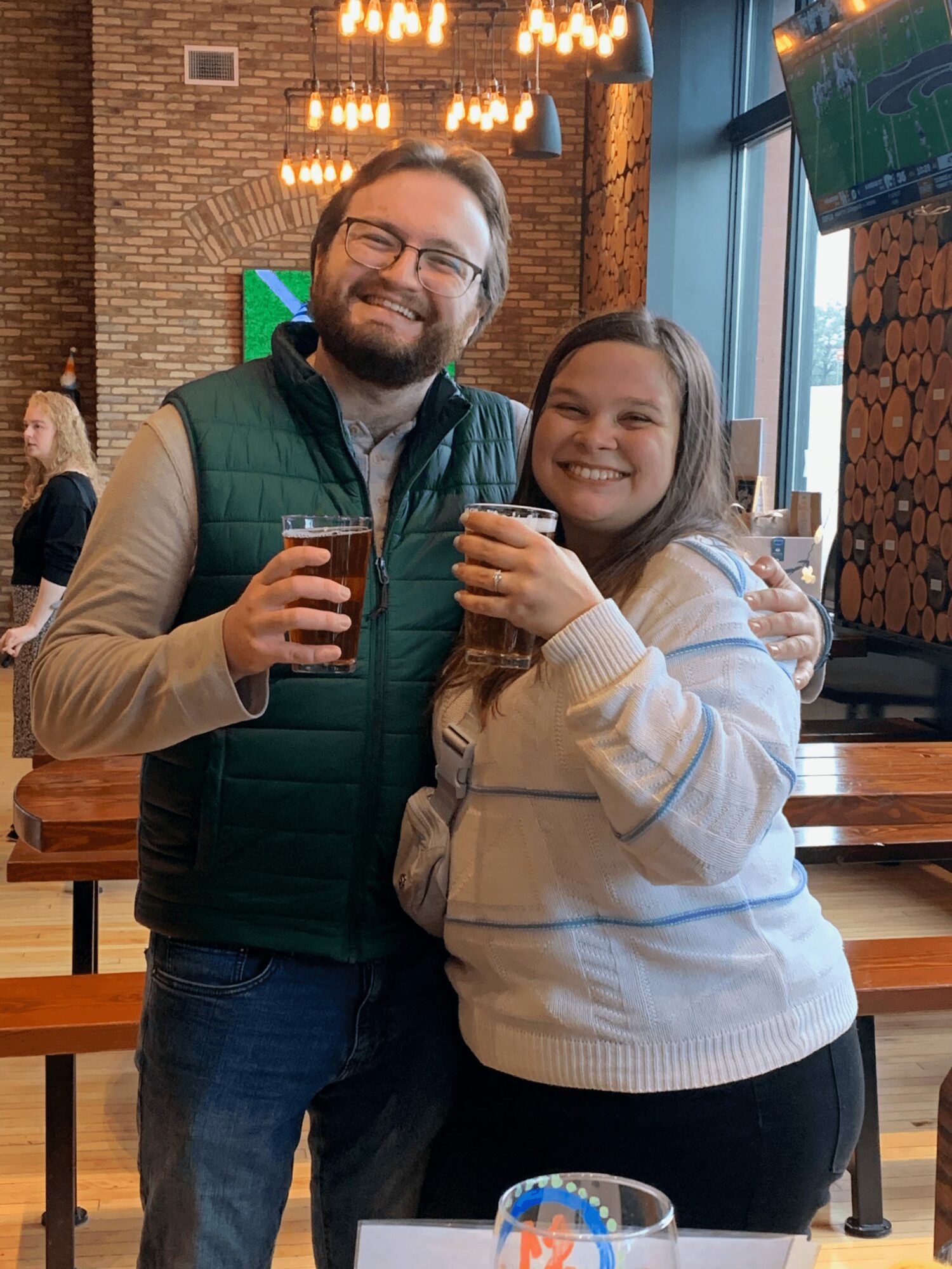Man and woman smiling, holding drinks, standing indoors with brick wall and wooden decor behind them.