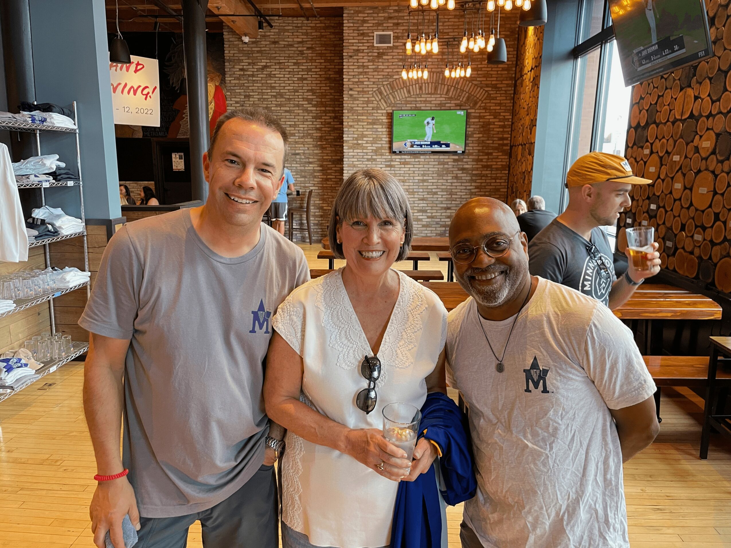 Three people smiling indoors, two men and one woman, with a bar and TV in the background.