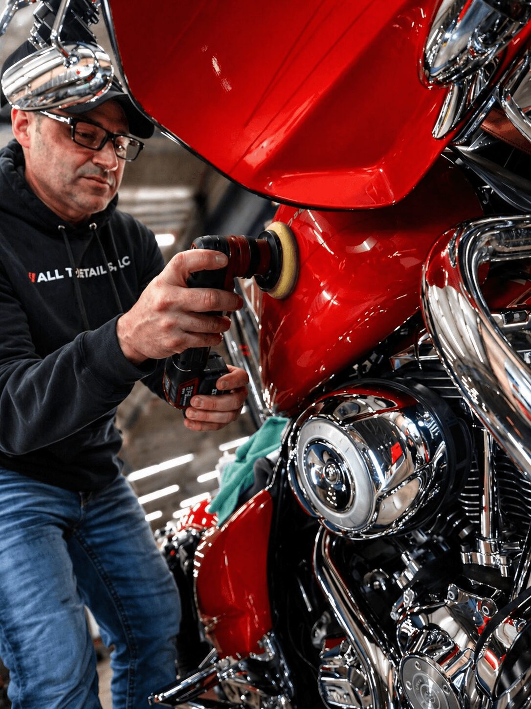 Person polishing a red motorcycle engine with a power tool, wearing glasses and a cap.