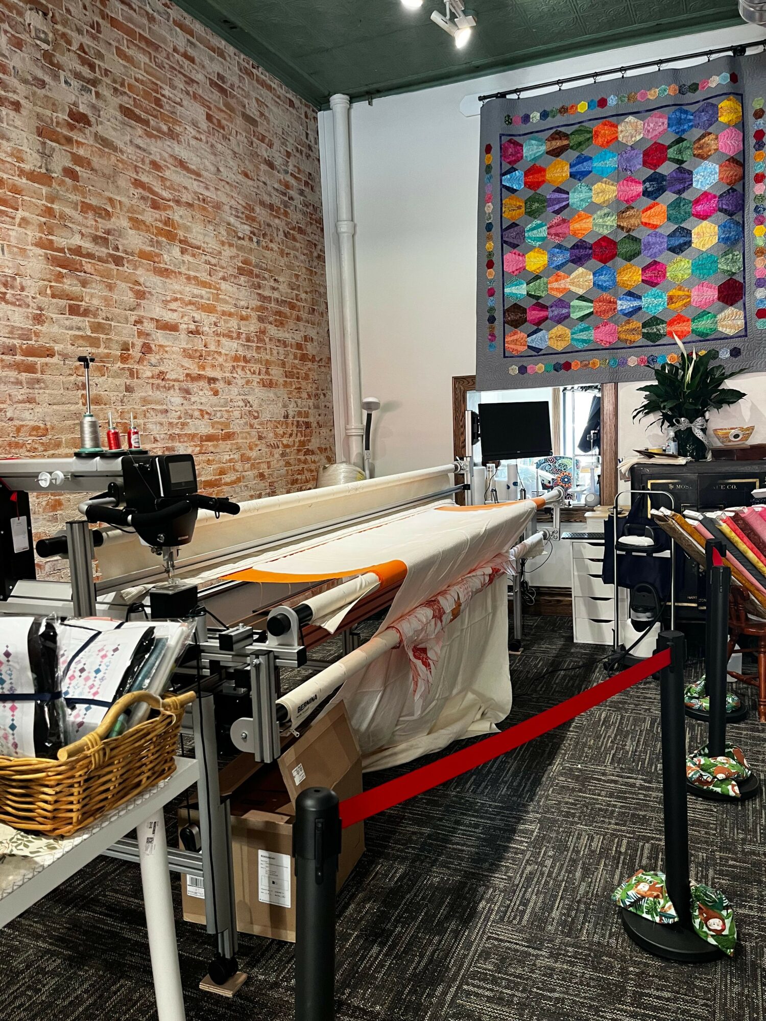 Office workspace with a colorful display, computer, and sewing machine on a table, brick wall, and carpeted floor.