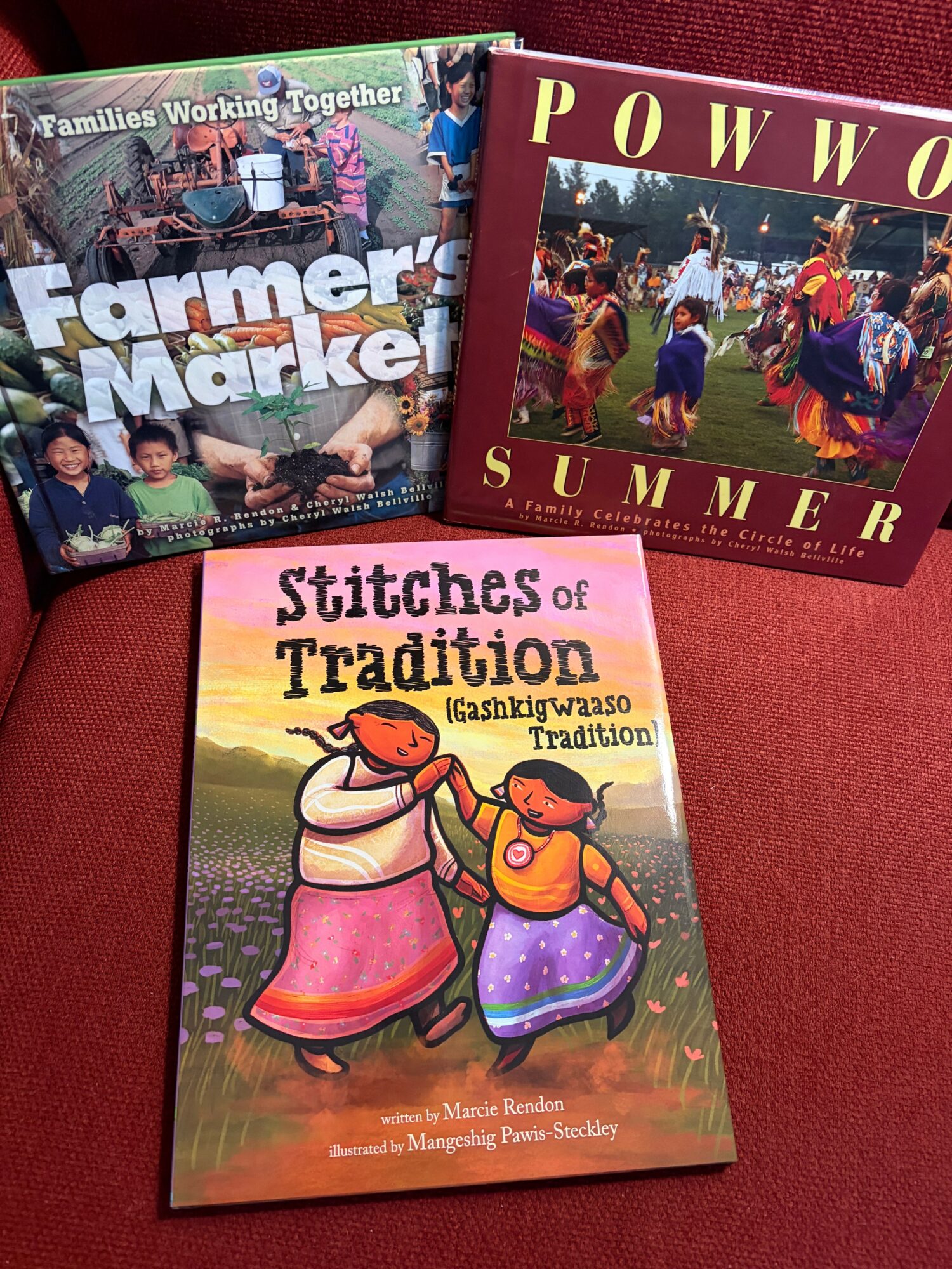 Three books on a red surface, with titles about farming, summer, and traditional stitches, featuring colorful covers and illustrations.