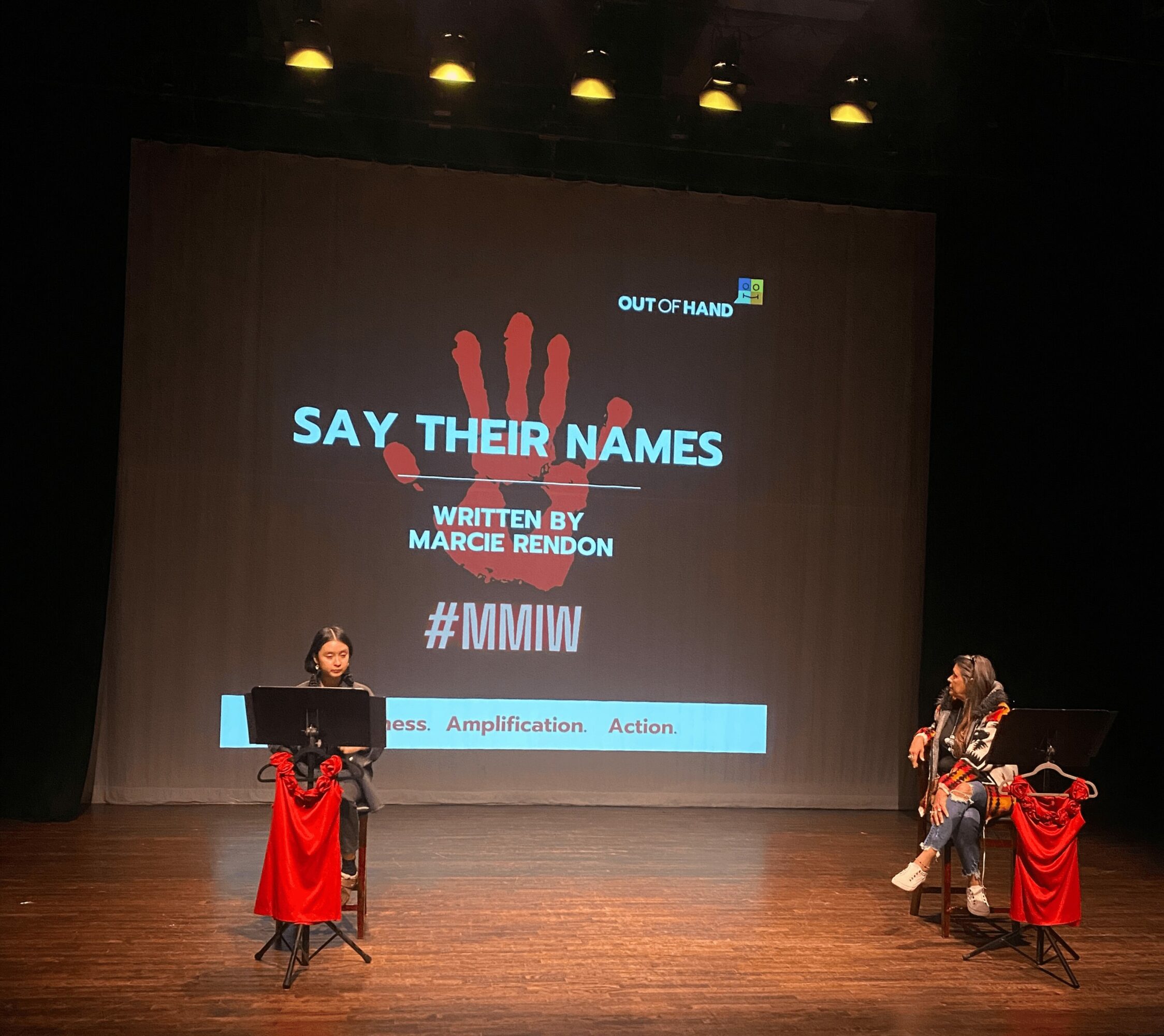 Two people sit on stage with a large screen behind them displaying a presentation titled 'Say Their Names' with a red handprint and hashtags.