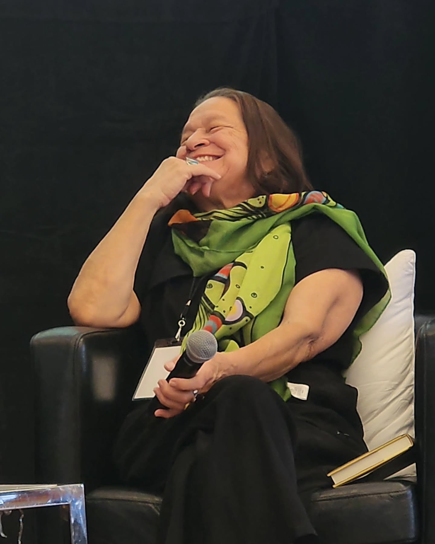 Woman sitting on a chair, smiling and holding a microphone, with a green scarf around her neck.