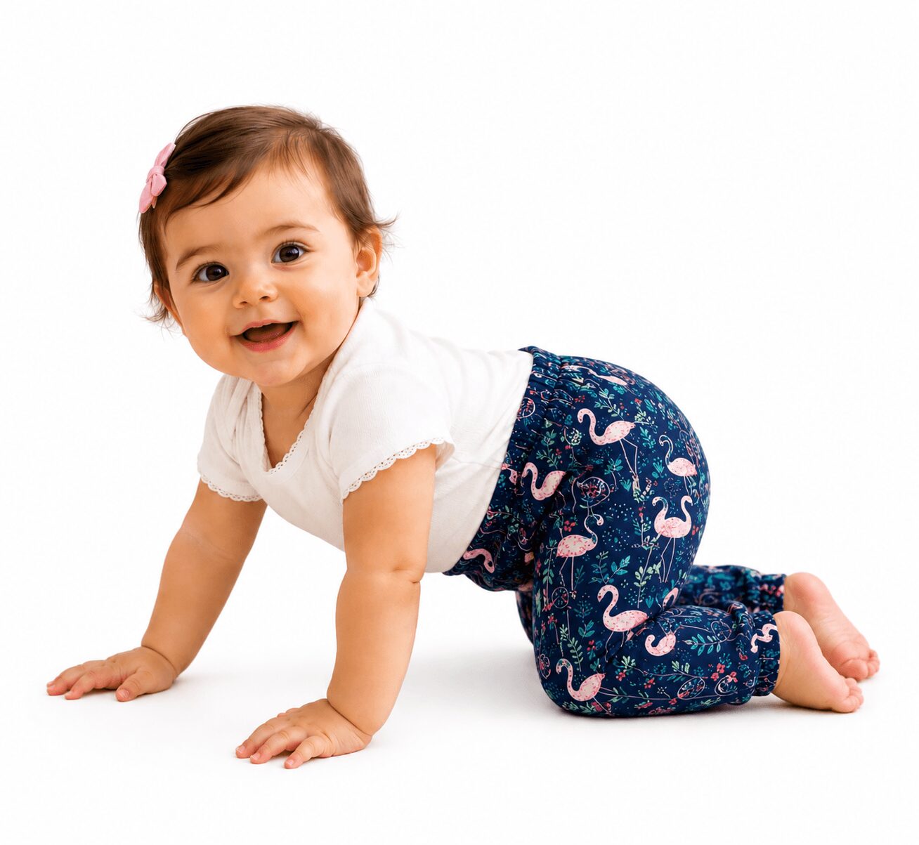 Baby crawling on white background, smiling, wearing a white shirt and patterned pants with flamingos, pink bow in hair.