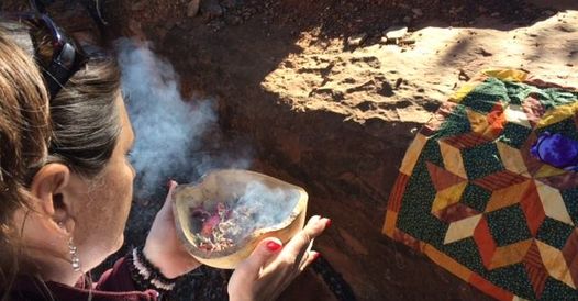 Person holding a bowl with smoke or steam, next to a colorful quilt on a dirt surface.