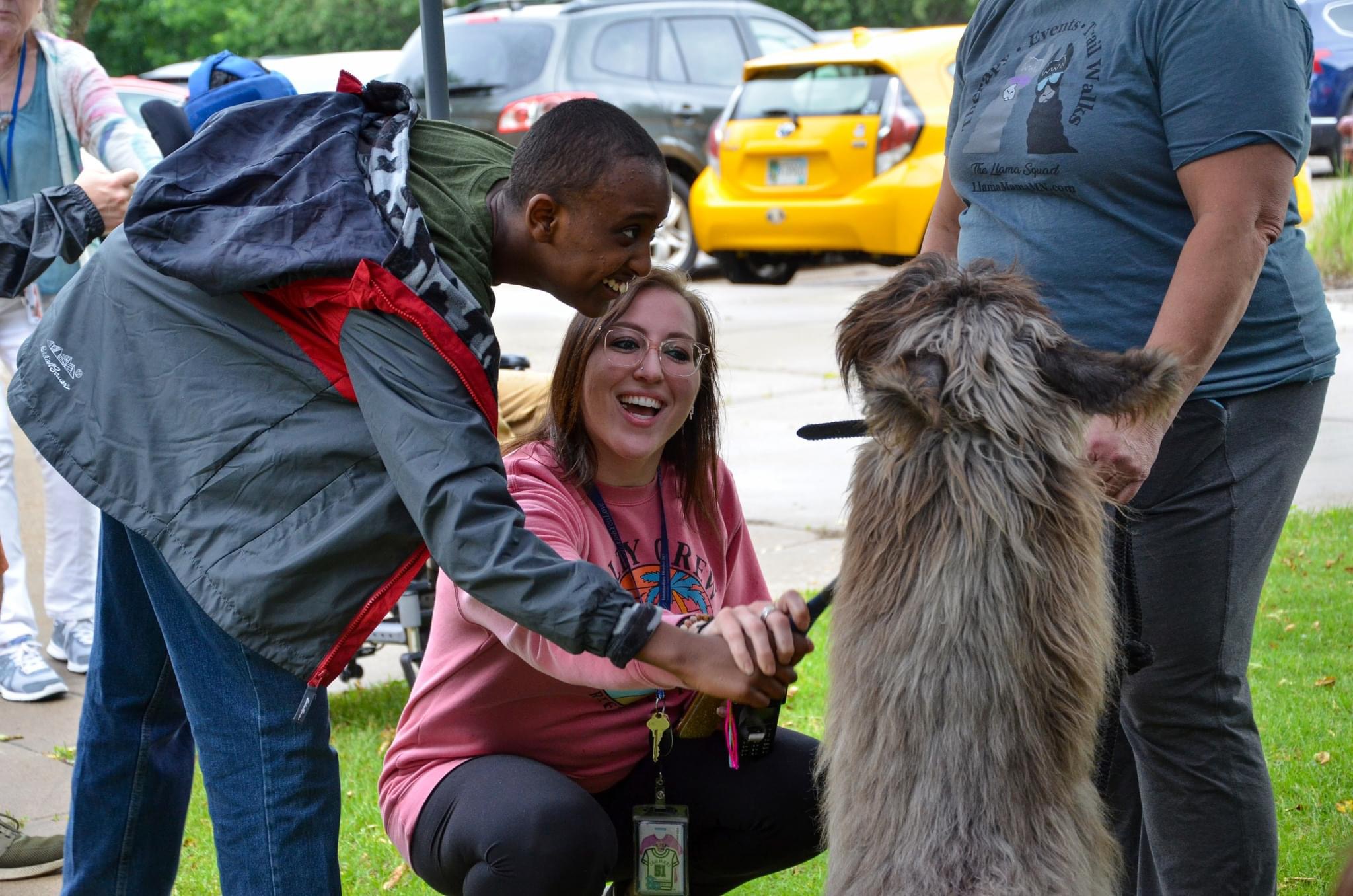 Person with backpack interacts with a large dog while a woman kneels nearby smiling, in an outdoor parking area.
