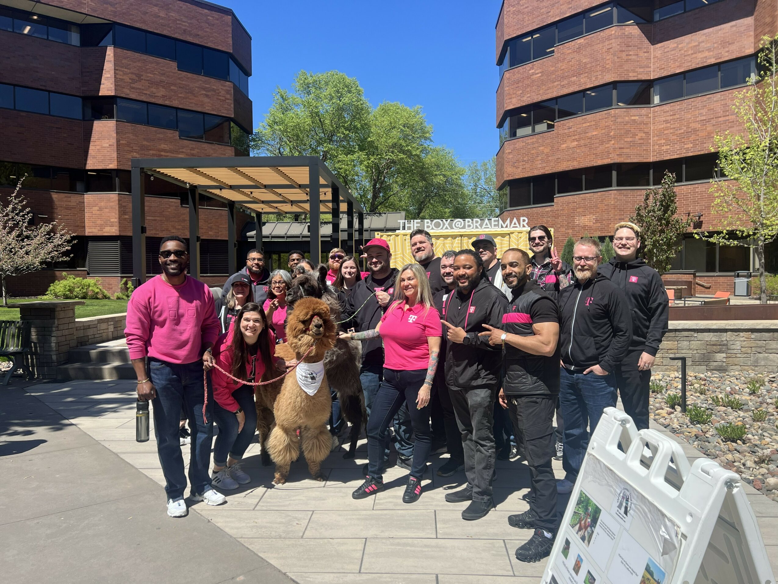 Group of people and a dog standing outdoors in front of modern buildings, smiling, with trees and a clear blue sky.