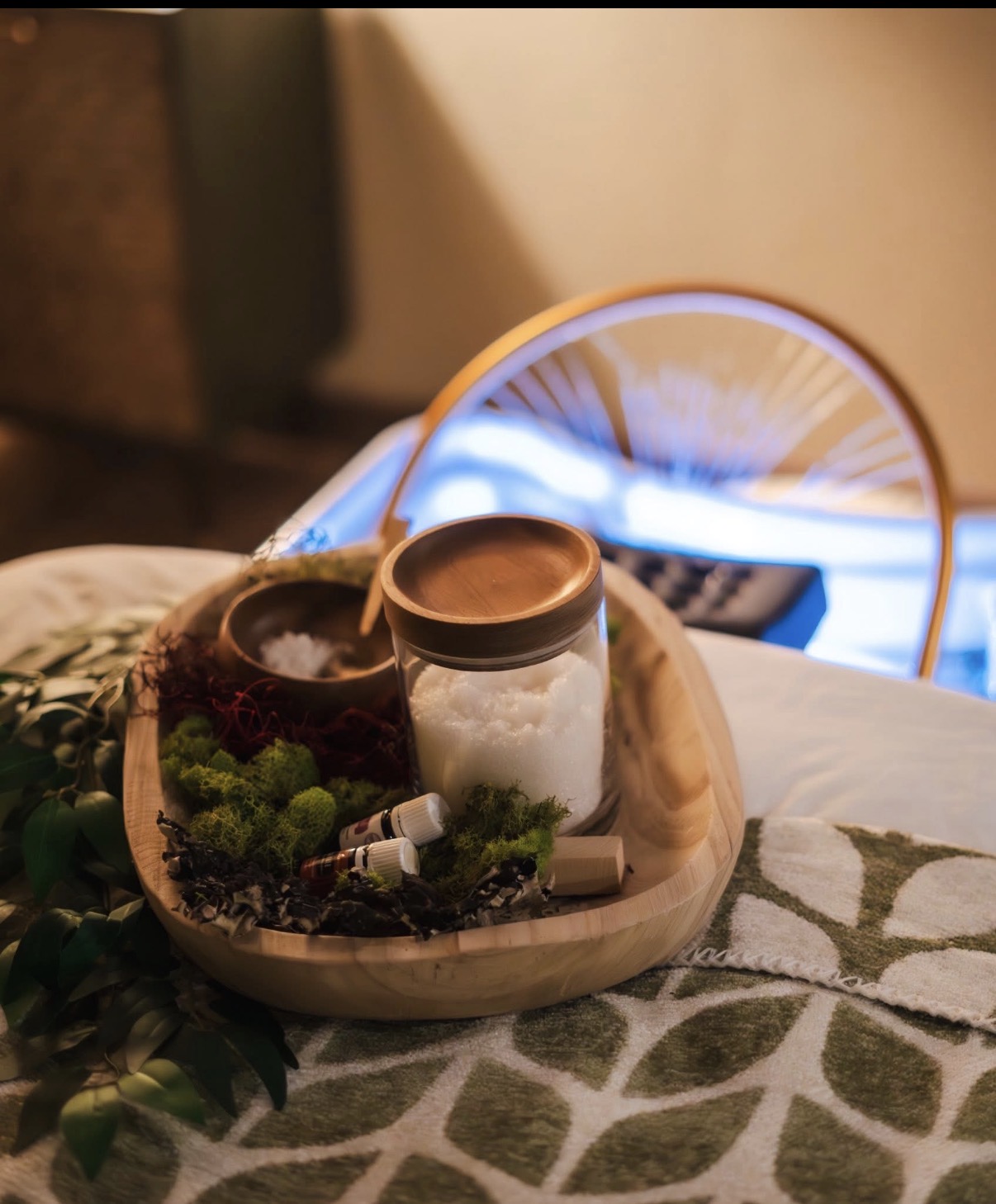 Tray with a jar of white substance, a small bowl, and green moss, on a bed with patterned sheets, in a cozy room.