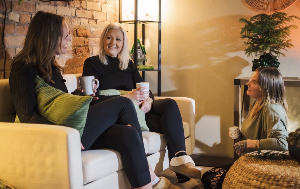 Three women and a girl sitting on a sofa and a chair, holding cups, in a cozy room with warm lighting.
