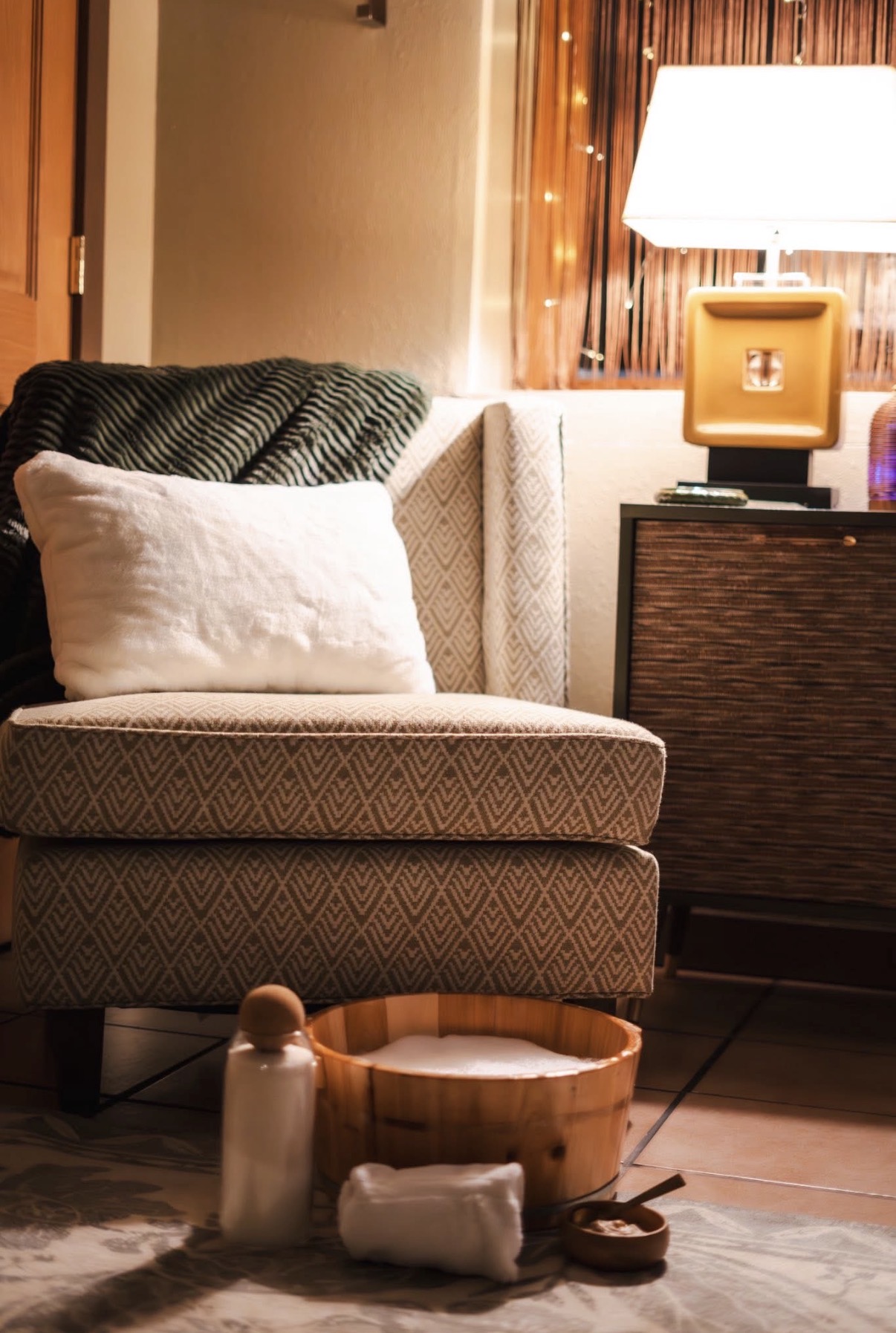 Living room corner with a patterned armchair, white pillow, wooden tray, and a side table with a lamp, warm lighting.