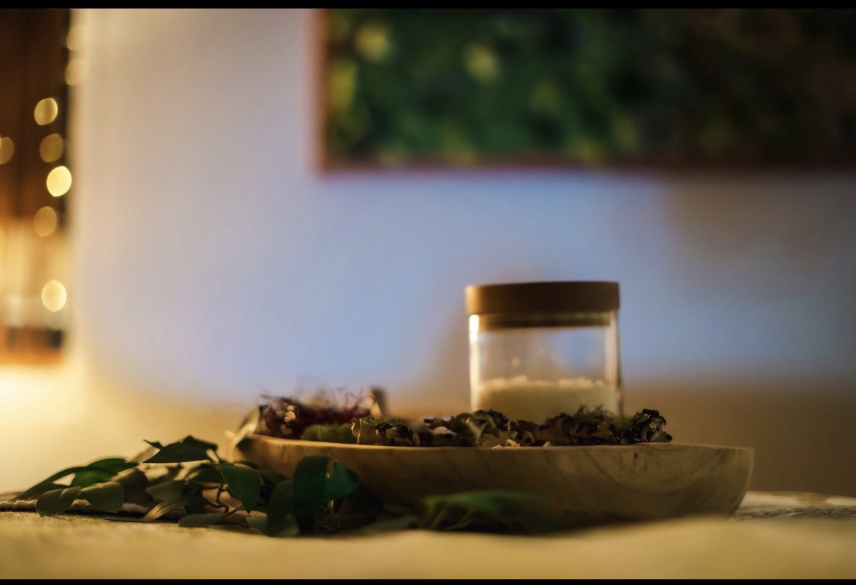 A glass jar on a wooden tray with dried leaves and flowers, indoors with blurred background and warm lighting.