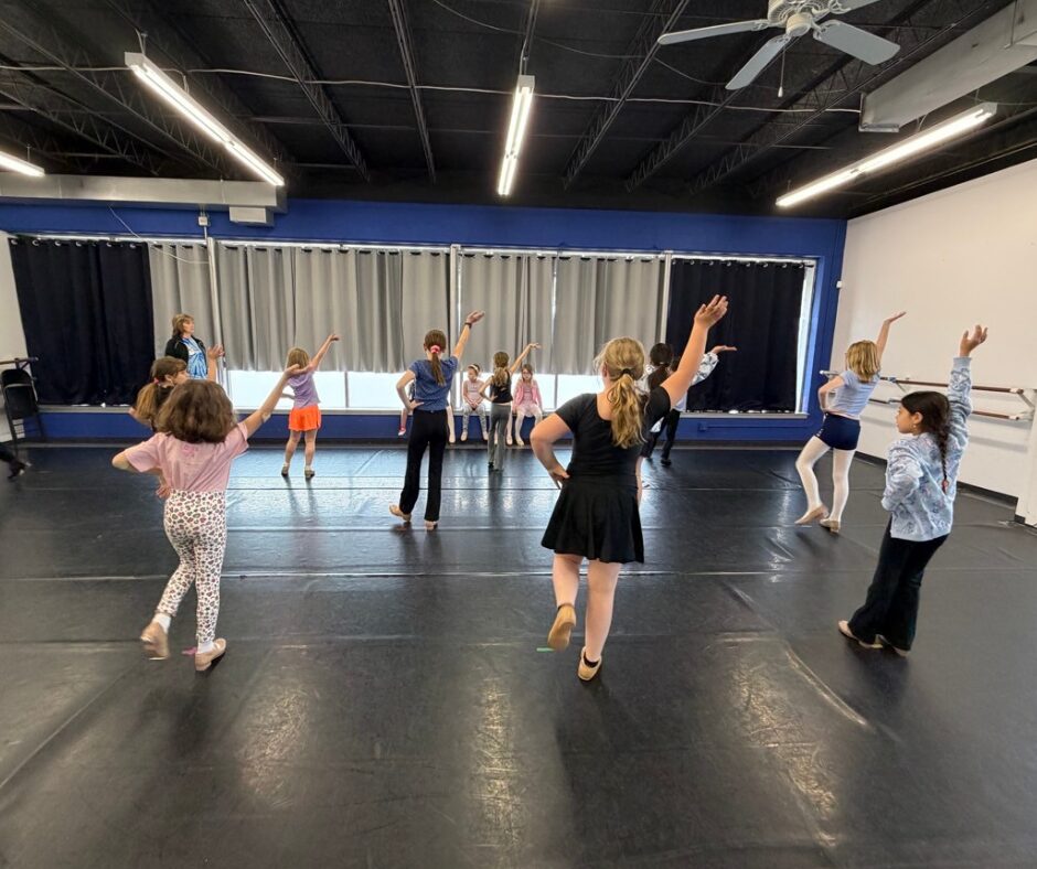 Children and adults dance in a spacious room with black flooring, blue and white walls, and ceiling lights.