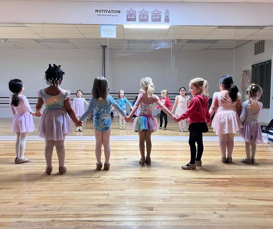 Children holding hands in a dance studio, facing a mirror, wearing colorful costumes and ballet shoes.