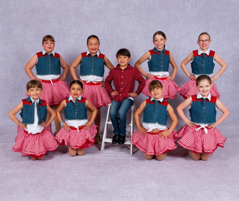 Group of nine children, eight girls in matching outfits and one boy in red shirt, posing against a gray background.