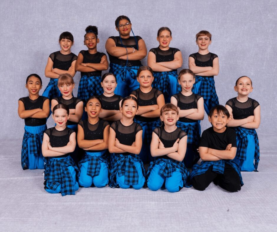 Group of 14 children in black tops and blue plaid skirts or pants, standing and kneeling, smiling, against a gray background.