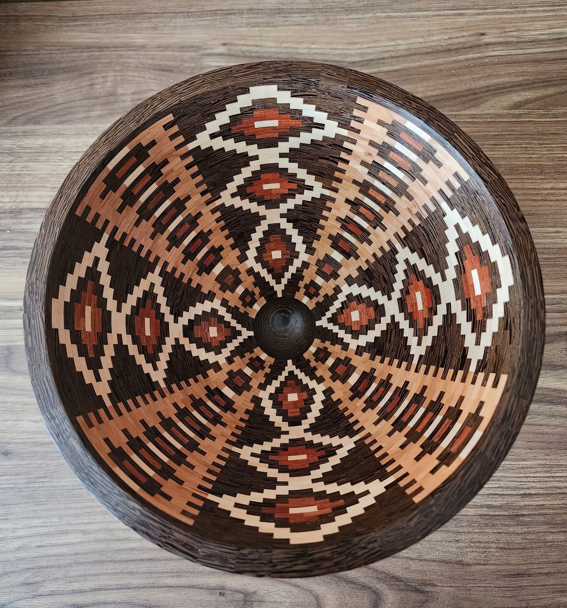 Decorative circular woven basket with geometric patterns in black, white, and reddish-brown, placed on a wooden surface.