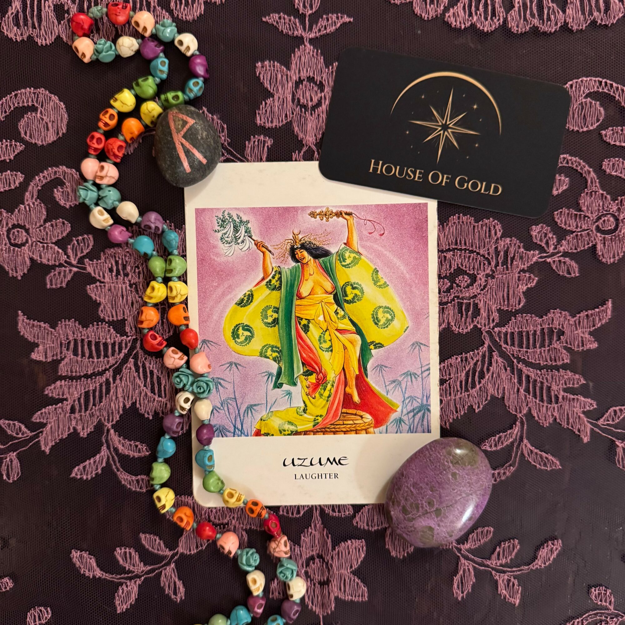 Colorful beaded necklace with a purple stone, card with a woman in traditional dress, and a black card with gold star and text, on purple patterned fabric.
