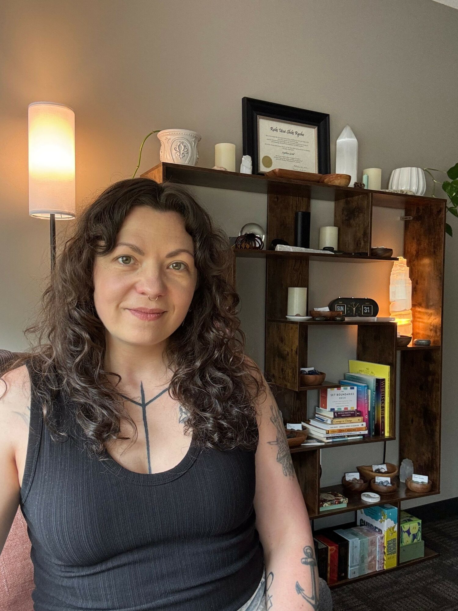 Woman with curly hair wearing a black top, smiling, in a room with bookshelves and a lamp.