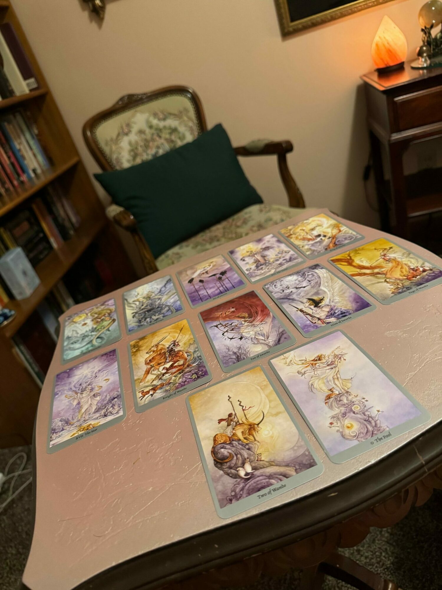 Table with tarot cards spread out, a chair with a cushion, and a bookshelf in the background.