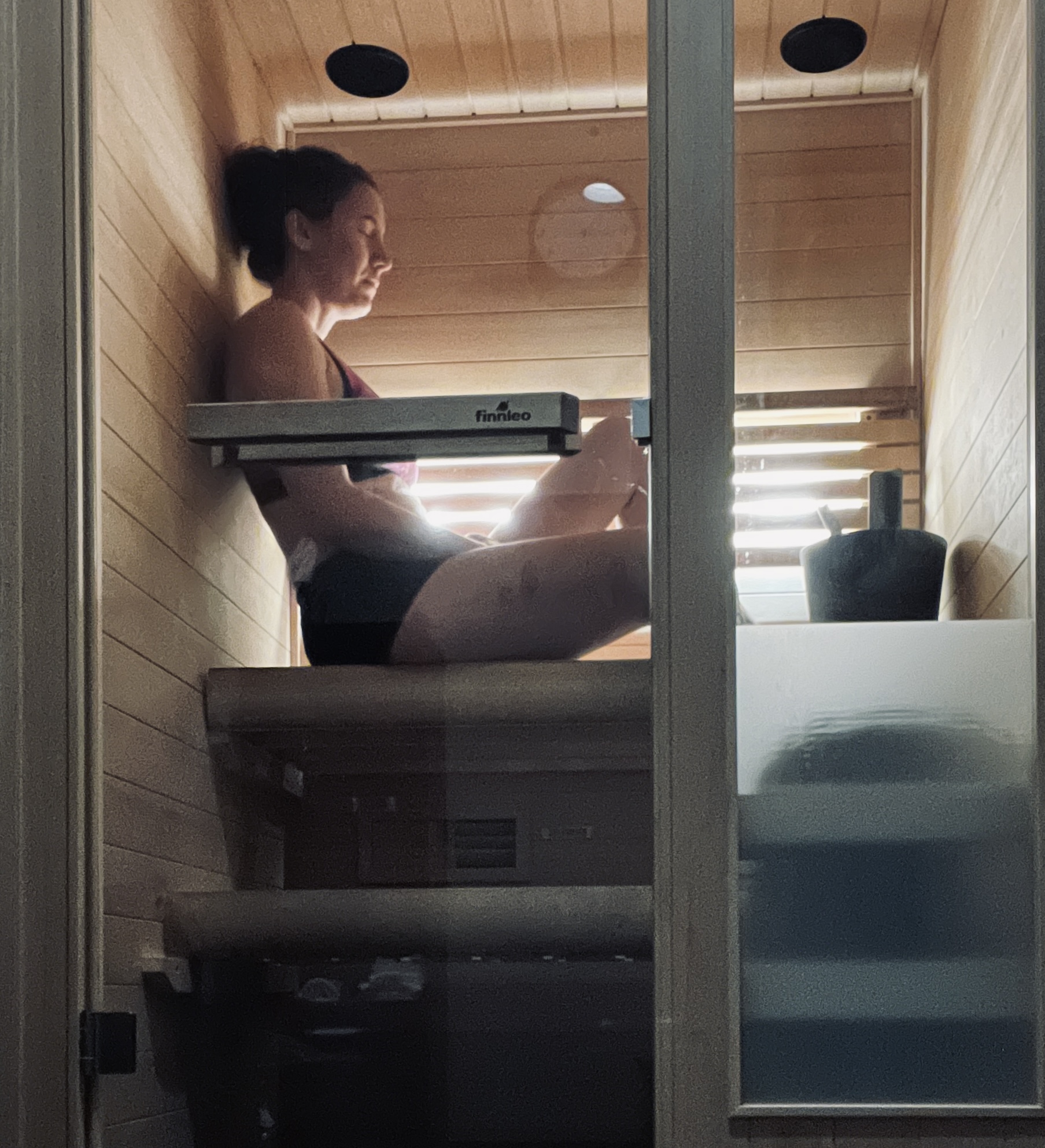 Person sitting inside a wooden sauna, facing a window with sunlight, with a bucket nearby.