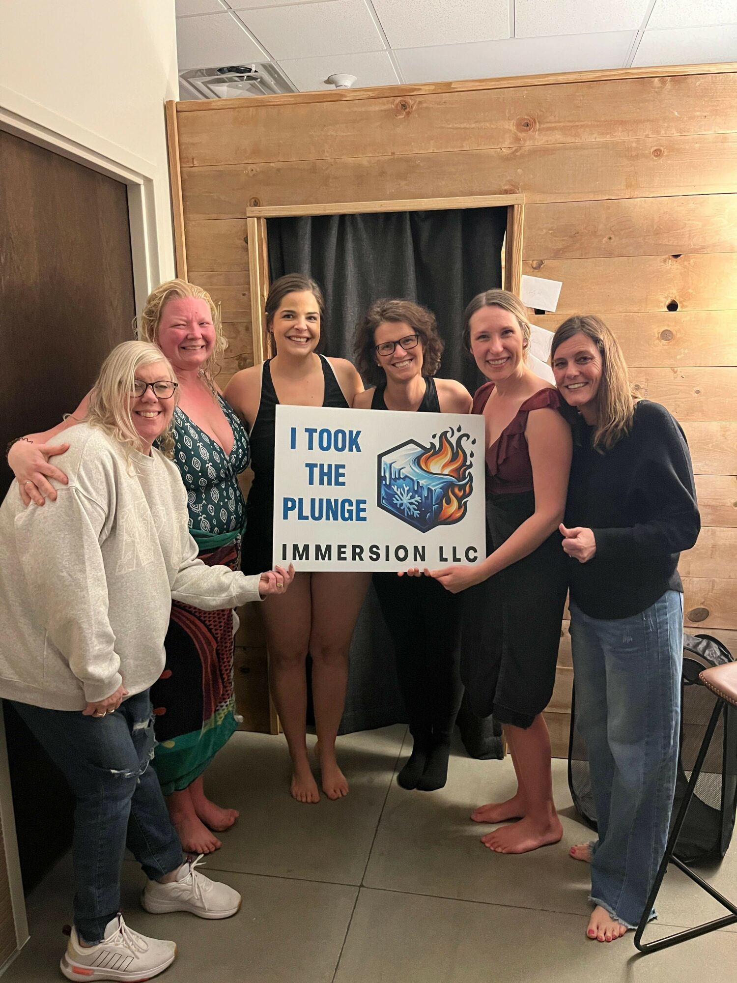 Four women and one girl holding a sign that says 'I took the plunge' in a room with wooden walls and a curtain.