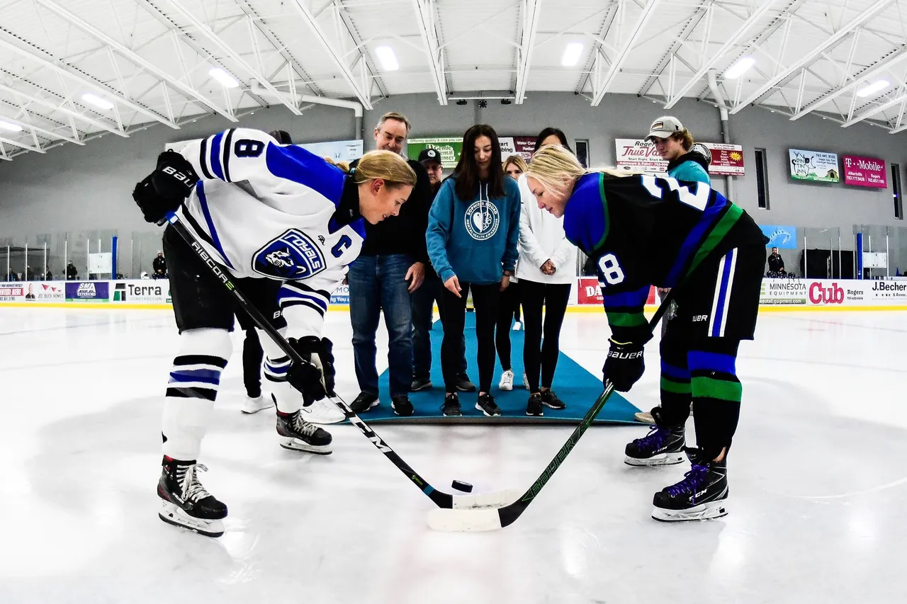 Hockey players and officials face off on ice rink, with spectators and scoreboard in background.