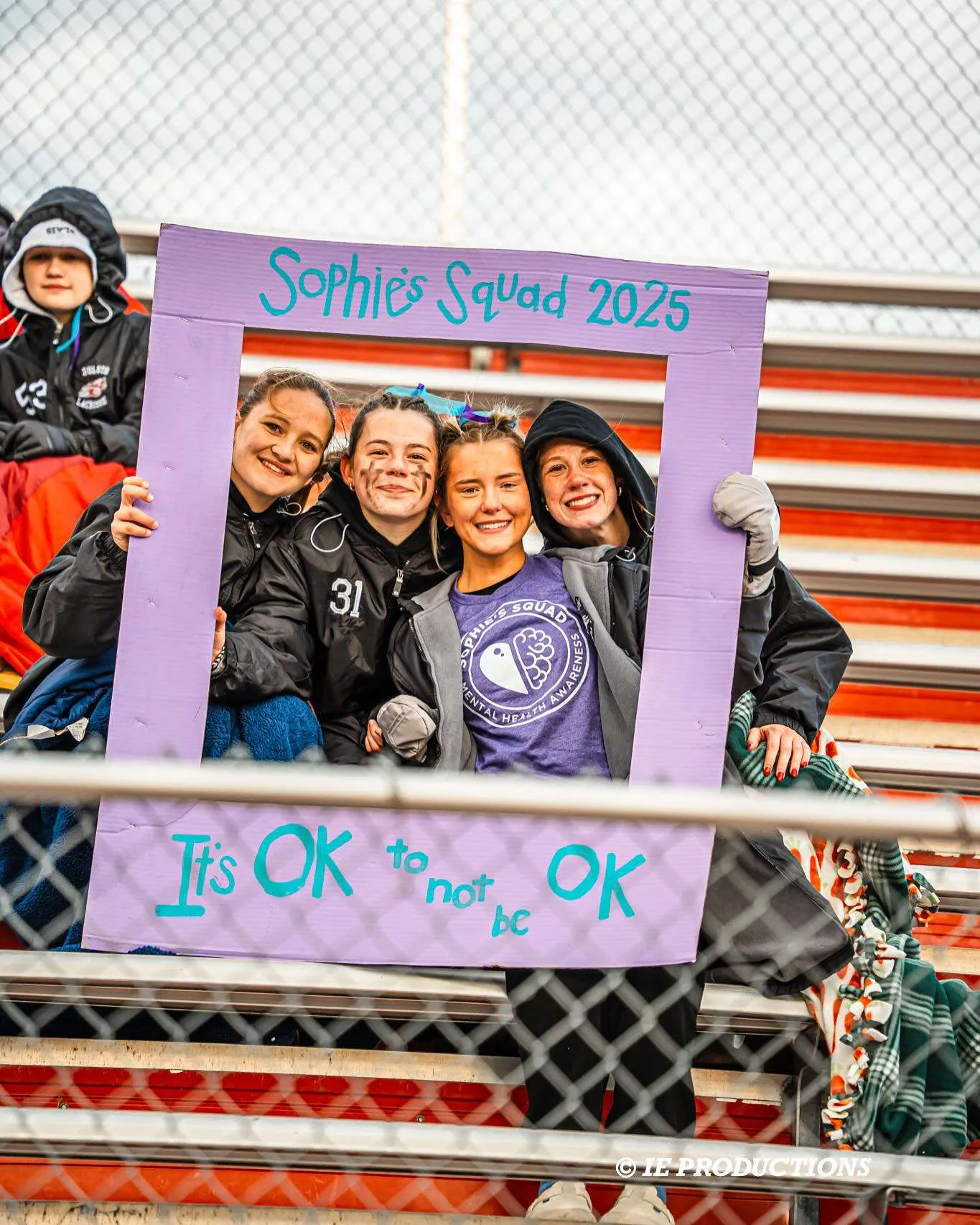 Four children smiling behind a purple frame with text, at a sports event, with a chain-link fence in foreground.