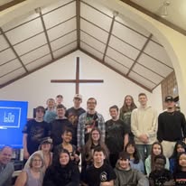 Group of people gathered inside a church with a cross on the wall behind them, some smiling and posing for the photo.