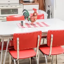 Kitchen table with red chairs, a rooster figurine on a checkered tablecloth, and kitchen appliances in the background.