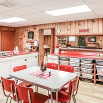 Dining area with a table, red chairs, and wooden wall paneling, featuring shelves and a checkered table runner.
