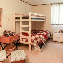 Bedroom with bunk bed, quilted bedspread, window with curtains, dresser, and chairs.