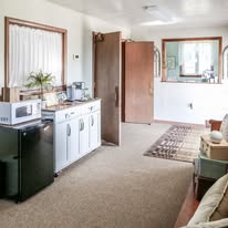 Kitchen with white cabinets, microwave, small refrigerator, open door, window with curtains, and a rug on beige carpet.