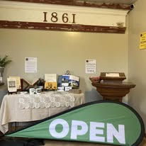 Interior with a table displaying framed photos and documents, a wooden stand, and a green 'OPEN' sign. The year 1861 is on the wall.