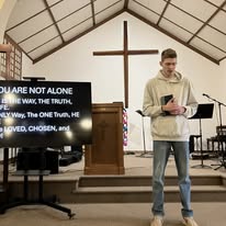 Young man reading from a phone in a church with a cross and a screen displaying text behind him.