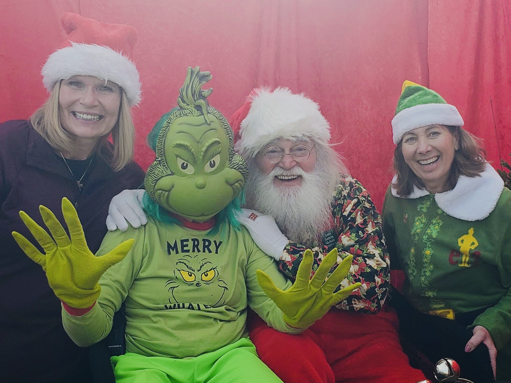 Four people, including a person in a Grinch costume, smiling in front of a pink background, wearing holiday attire.