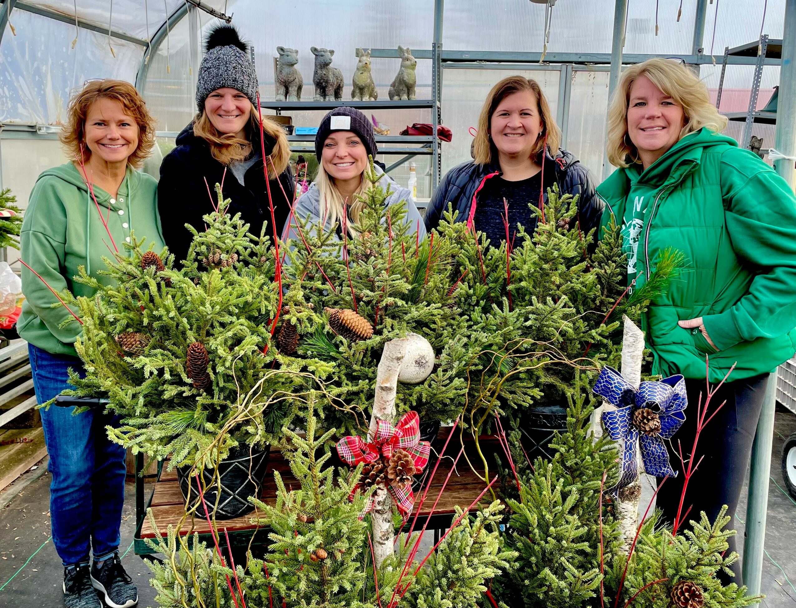 Five women stand behind a large decorated Christmas wreath display inside a greenhouse, smiling at the camera.