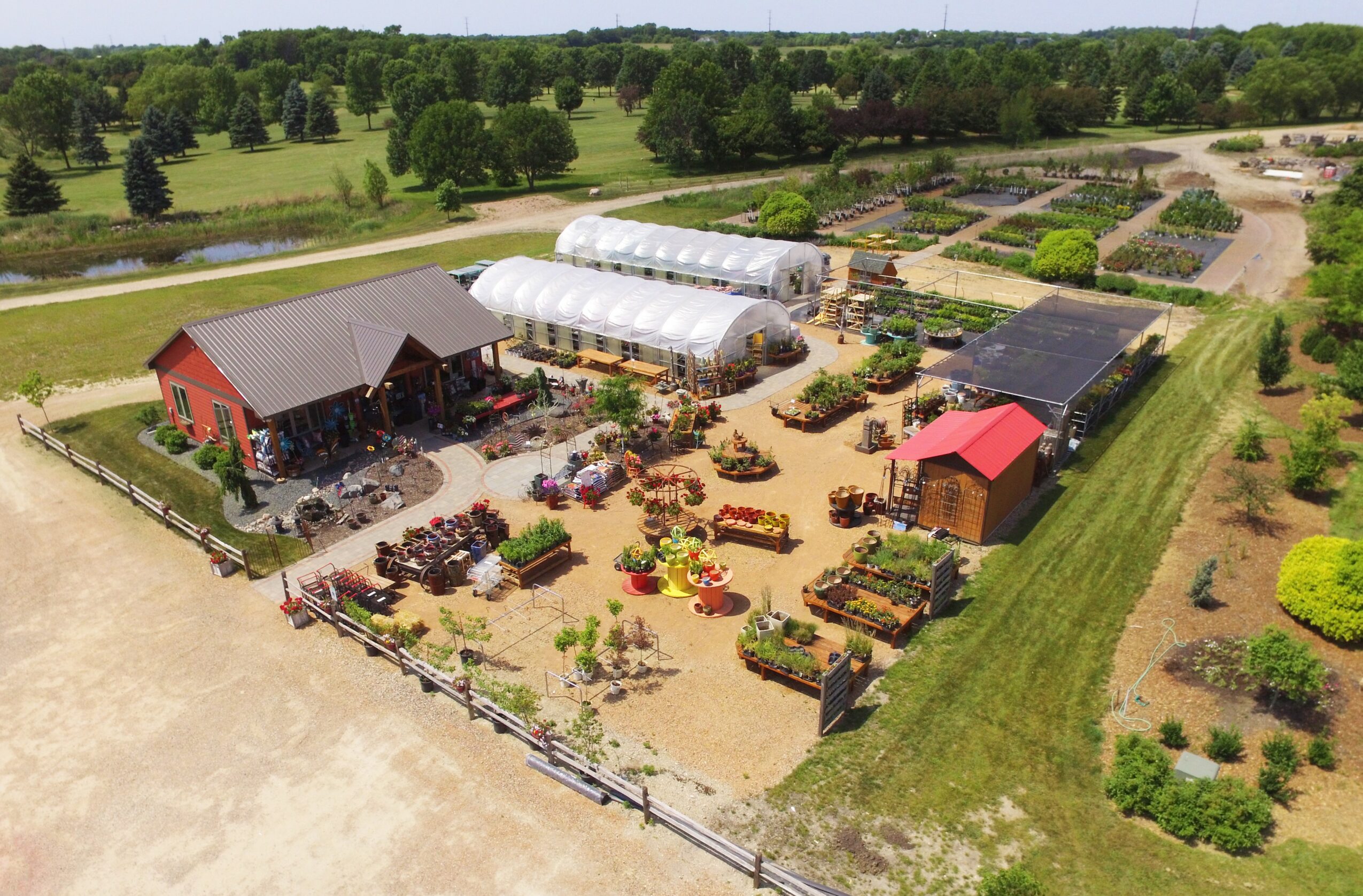 A farm with greenhouses, barns, and garden beds, surrounded by trees and open fields, viewed from above.