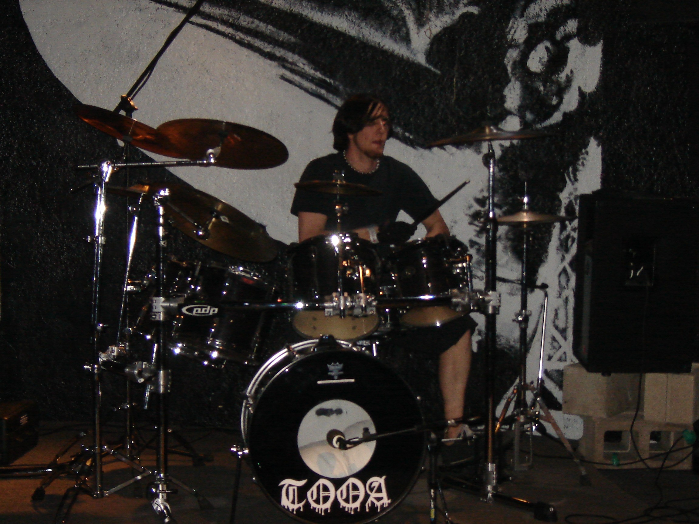 Person playing drums on stage with a black and white backdrop, surrounded by musical equipment.