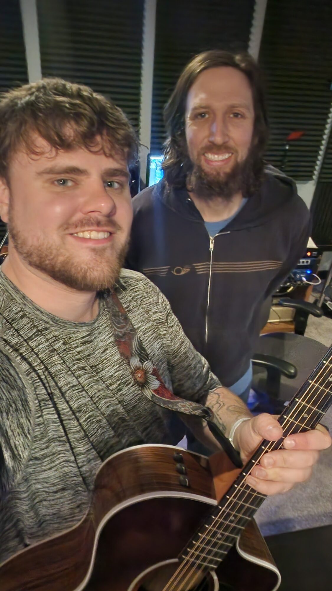 Two men smiling, one holding a guitar, in a room with soundproofing panels and equipment.