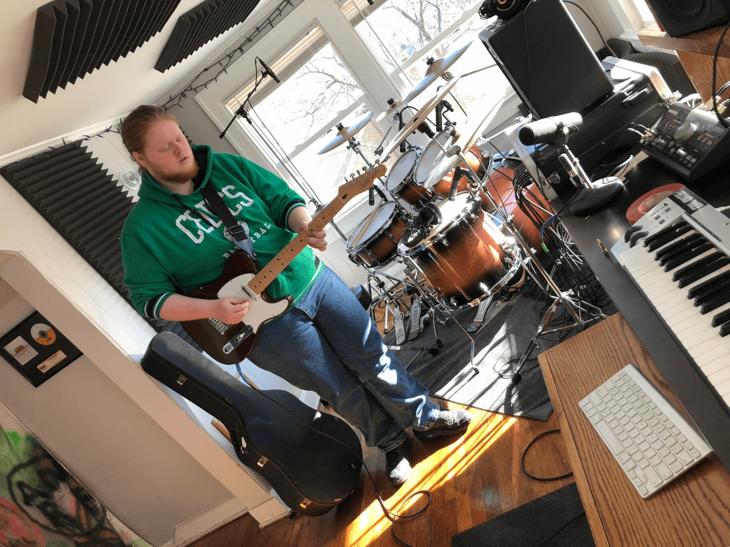 Young person playing electric guitar in a music studio with drums, keyboard, and soundproofing panels.