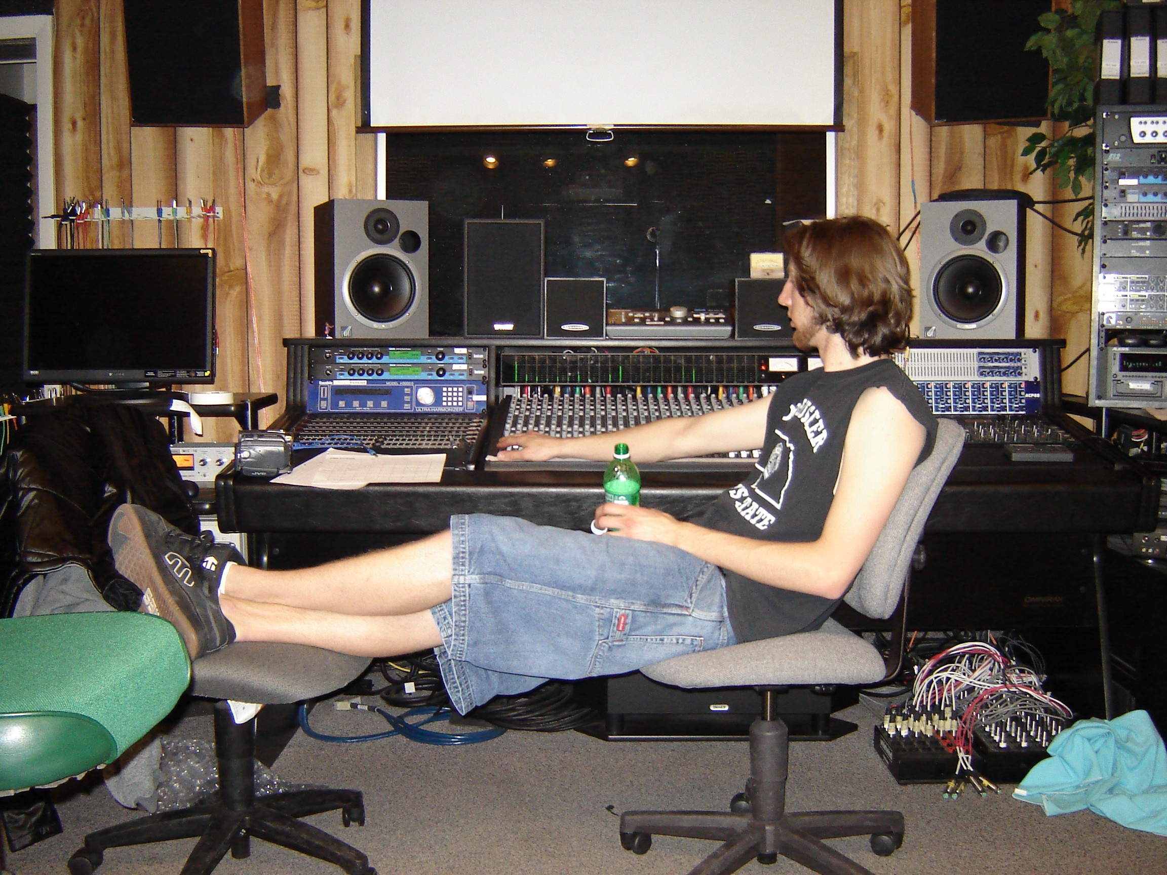 Person sitting in a recording studio with equipment, holding a water bottle, with legs stretched out on a chair.