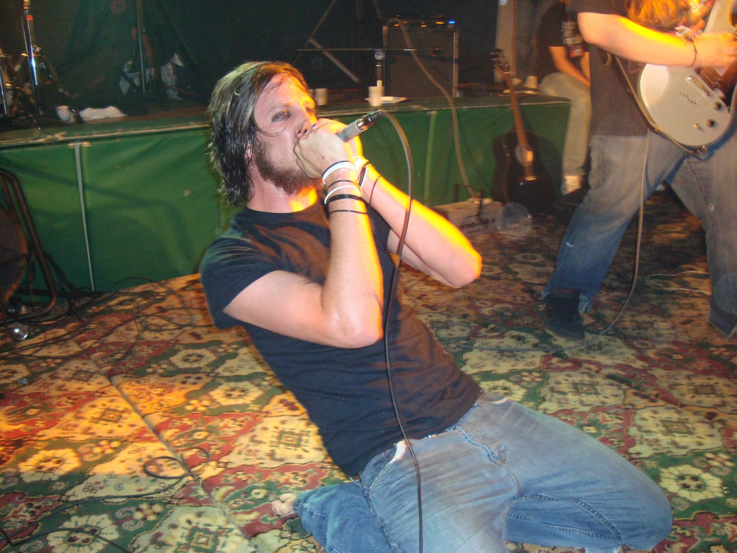 Person with long hair and a beard singing into a microphone on stage, wearing a black t-shirt and jeans, with a patterned carpet beneath.