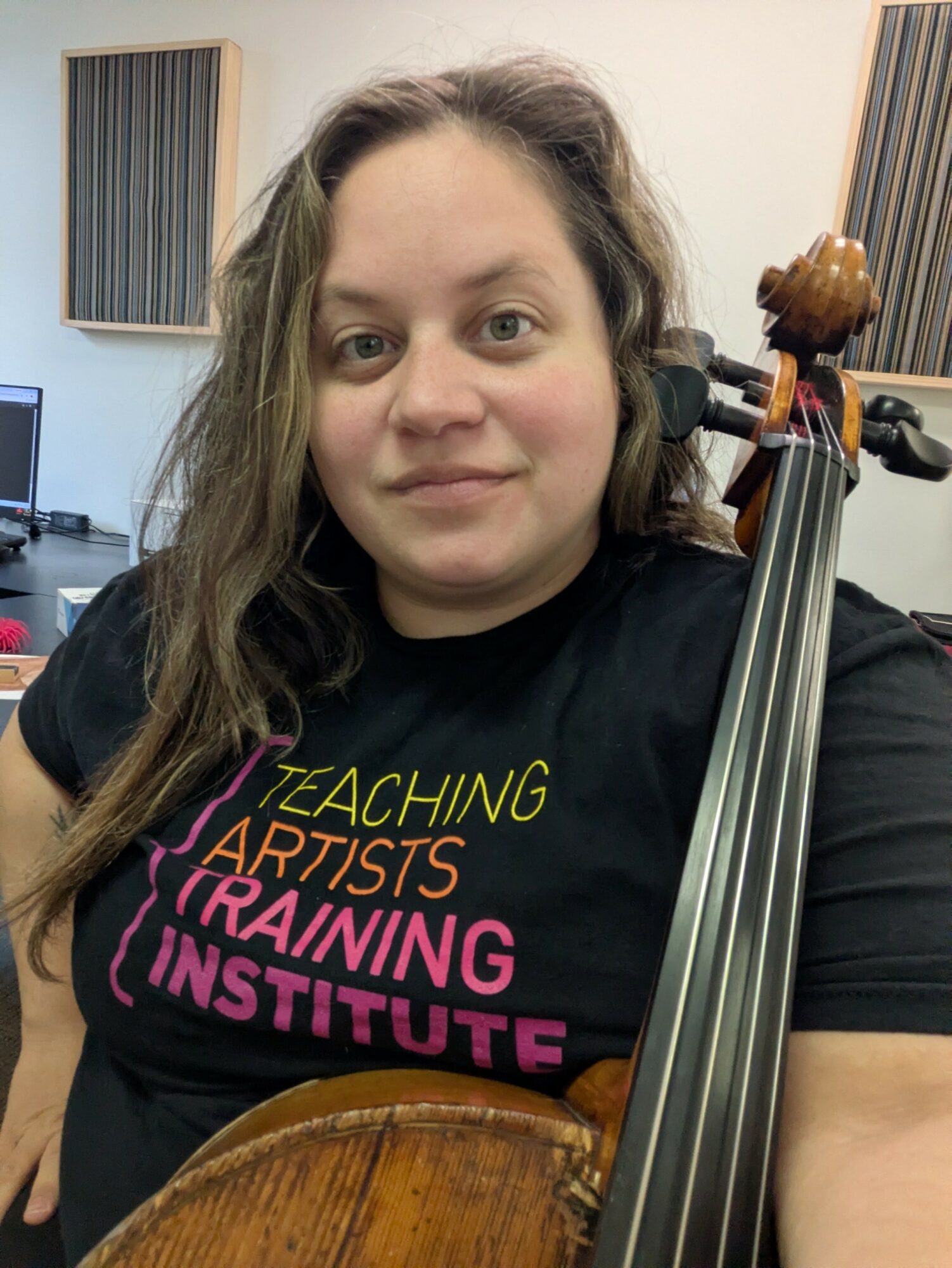 Person with long hair holding a string instrument, wearing a black T-shirt with colorful text, in a music studio.
